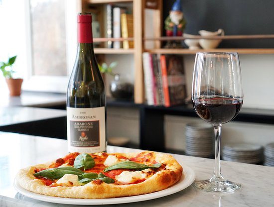 A cozy culinary scene featuring a freshly baked pizza with buffalo mozzarella, fresh basil, and cherry tomatoes, accompanied by a glass of red wine and a bottle of Amarone wine on a marble countertop. Perfect for a relaxed dining experience at home. 