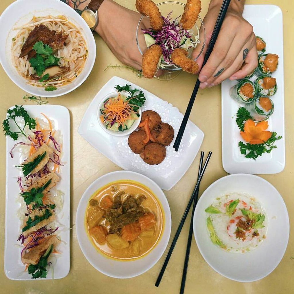 An overhead view of a diverse Asian feast featuring multiple dishes: a bowl of Pho with tender slices of beef, vibrant sushi rolls with fresh salmon, crispy spring rolls accompanied by a tangy dipping sauce, rich curry with chunks of meat and vegetables, deep-fried fish cakes, and a fresh noodle salad, all arranged neatly on a table, ready to be enjoyed.