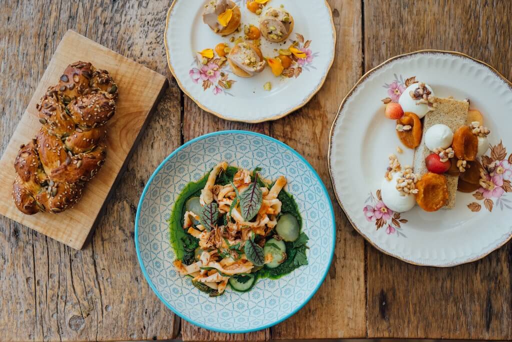 Assorted gourmet dishes beautifully presented on rustic wooden table, featuring a seed-topped braided bread on a wooden board, a vibrant salad with shredded chicken and fresh greens on a blue patterned plate, and a dessert plate with various fruits and cream on a vintage floral plate. A visual feast that captures the essence of fine dining.