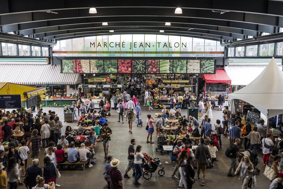 A bustling scene at Marché Jean-Talon in Montreal, one of the largest public markets in North America. The image captures a lively atmosphere with diverse crowds of people shopping, socializing, and enjoying a variety of fresh produce and local goods under the market's iconic signage.
