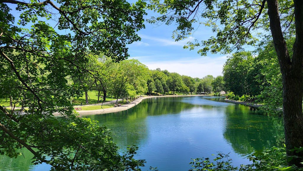 A serene view of Parc La Fontaine in Montreal, showcasing a tranquil lake surrounded by lush greenery and a neatly maintained pathway. The perspective through the trees offers a peaceful retreat in one of the city's most beloved parks, ideal for strolls, cycling, or just enjoying the outdoors
