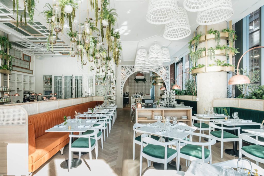 Elegant and stylish restaurant interior decorated with an abundance of greenery, featuring white tables and green velvet chairs under unique white lattice chandeliers. The space is filled with natural light, enhancing the botanical atmosphere and sophisticated layout of the dining area.