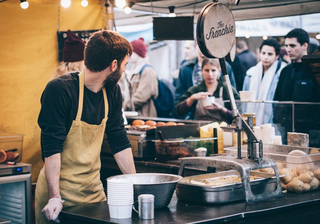 A vendor in an apron serving food at a market stall, with customers in the background, perfect for a casual and fun bachelorette party outing.